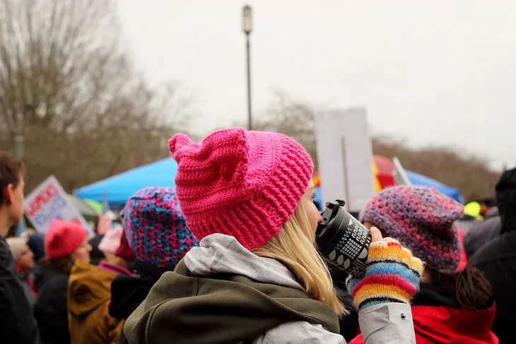 A blonde woman wearing a pink hat drinking a coffee, at the women’s march.