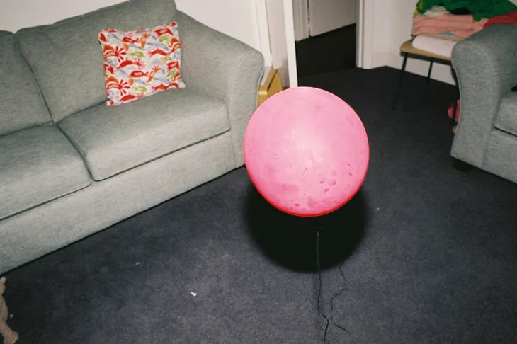 A pink helium balloon floats in the middle of a living room, low to the ground