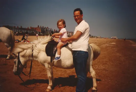 A three-year-old girl is sitting on a saddle on a donkey while a man holds her in place. They are on a beach during summer.