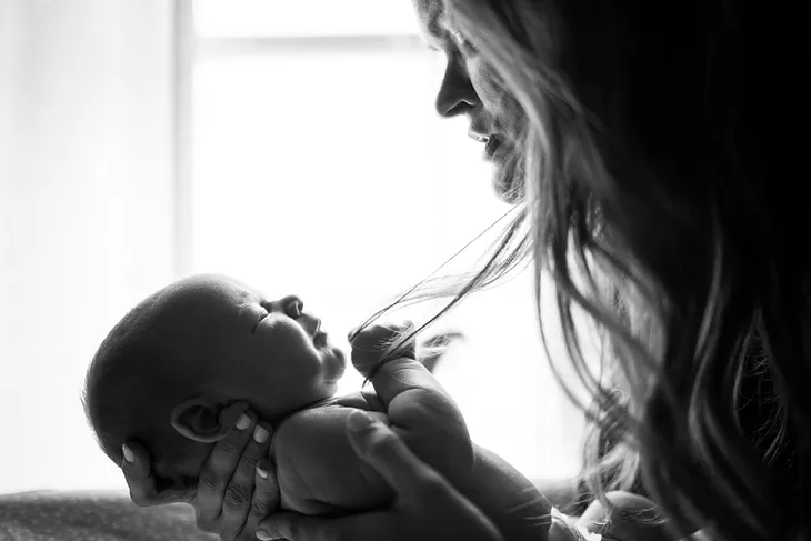 Black-and-white photo of a baby facing a mother in profile, in front of a window. The baby has hold of a strand of the mother’s hair and has closed eyes. The mother is looking intently at the baby.