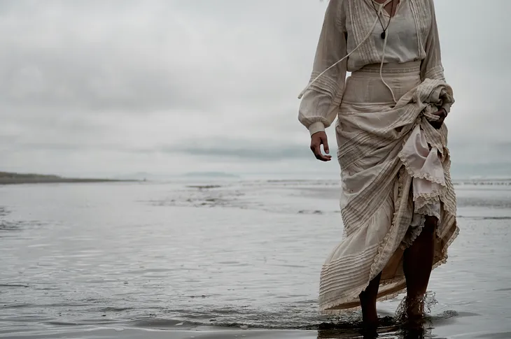 A woman in a white dress walks through the shallows along a beach, the hem of her dress getting wetter as she goes.