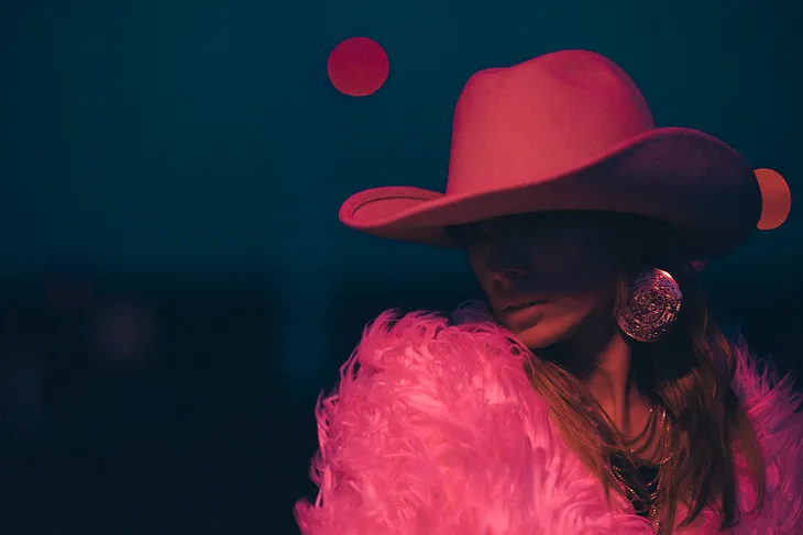 A woman wearing a bright pink cowboy hat and a bring pink feather jacket. The woman has long hair and her face is obscured by the shadow from the hat. The background to the picture is dark except for a bright pink moon.