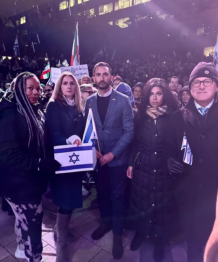 Stephen Lecce stands with a group of people at an evening event, holding Israeli flags, with a crowd illuminated by ambient light in the background, indicating a solemn gathering.