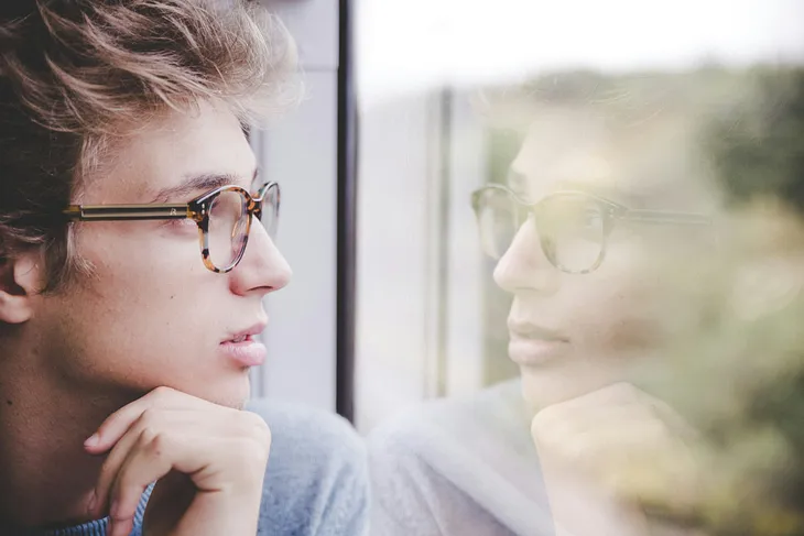 young man with glasses looking out the window, in which we see his reflection.