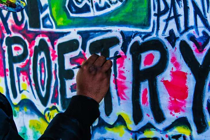 a man’s hand using a brush to finish painting the word ‘poetry’ in a colourful graffiti style
