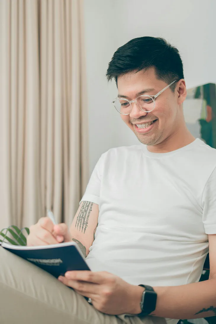 A photo of a smiling Asian man smiling while gladly scribbling something down in a notebook.