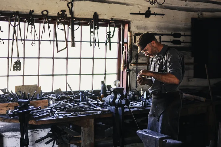 Man wearing leather apron in front of a window. Numerous tools on bench behind him and over the window.