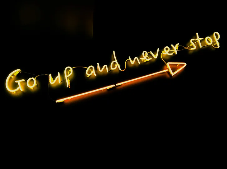 Neon sign of an arrow pointing up with the words: “Go up and never stop”