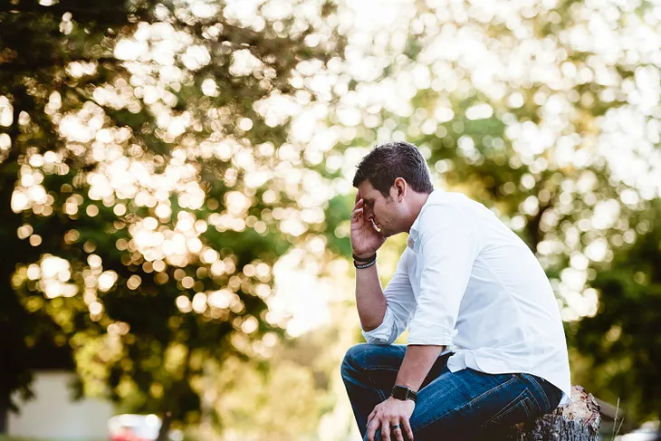 A man sitting outside on a bright day with his hand touching his forehead and his eyes closed as if trying to maintain composure during a difficult time. This is demonstrative of a mans attempts to hide his feelings or feeling the need to go through tough emotions alone.