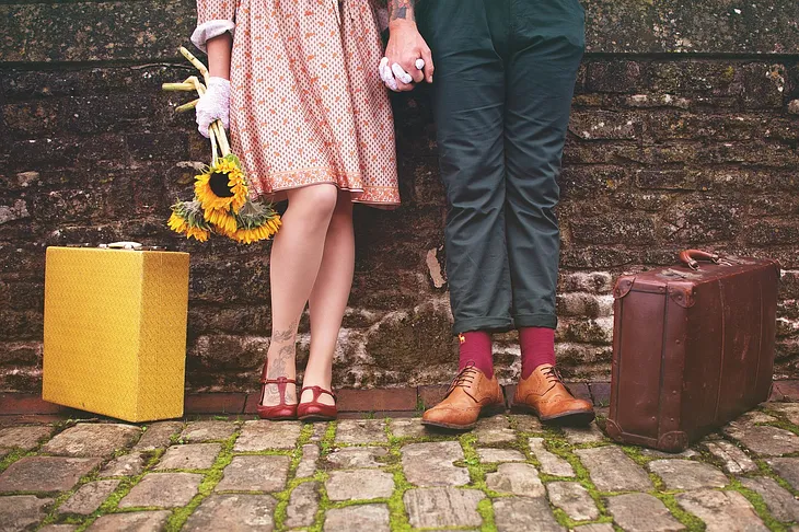 Light-skinned woman holding a bunch of sunflowers, wearing a red and white dress, white gloves, and red dressy shoes, standing next to a yellow vintage suitcase, holding hands with a light-skinned masculine-looking person wearing navy blue pants, red socks, and sable short boots, standing next to a brown vintage suitcase. They both have visible tattoos on ankle and wrist, and are standing against a brown brick wall, on white cobblestones with moss in the cracks.