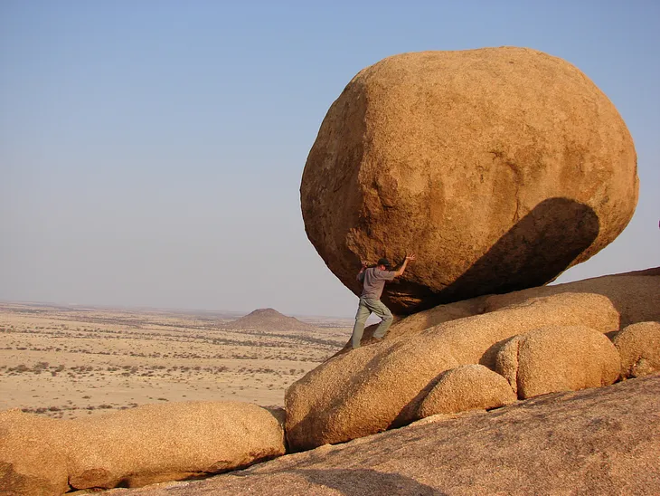 A man in a desert looking like he’s holding a big rock