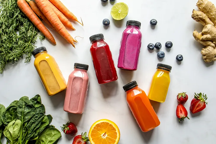 Many bottles of smoothies of different colors, surrounded by fresh fruit.