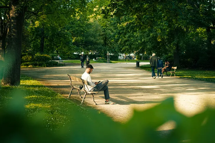 A tree-lined street in a park, a man sitting on a bench with his phone, and a few people strolling.