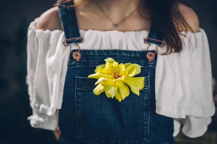 A woman wearing overalls with a yellow blossom tucked inside her bib pocket.