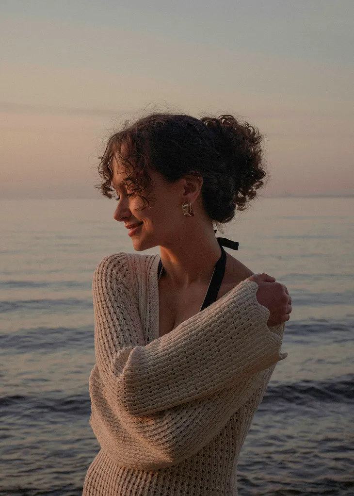 A woman with curly hair standing on a sea shore with her right arm on her left shoulder