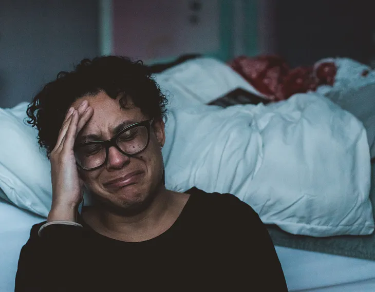 A woman sitting on the floor beside her bed, hand on her head, looking sad and depressed.