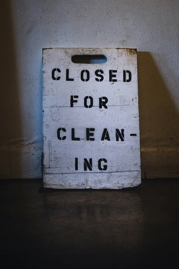 Picture of a an old, dirty, white sign that says closed for cleaning leaning against a wall in a slightly dark room. Cleaning has an apostrophe in between the first n and the only i, so that’s why I included it.