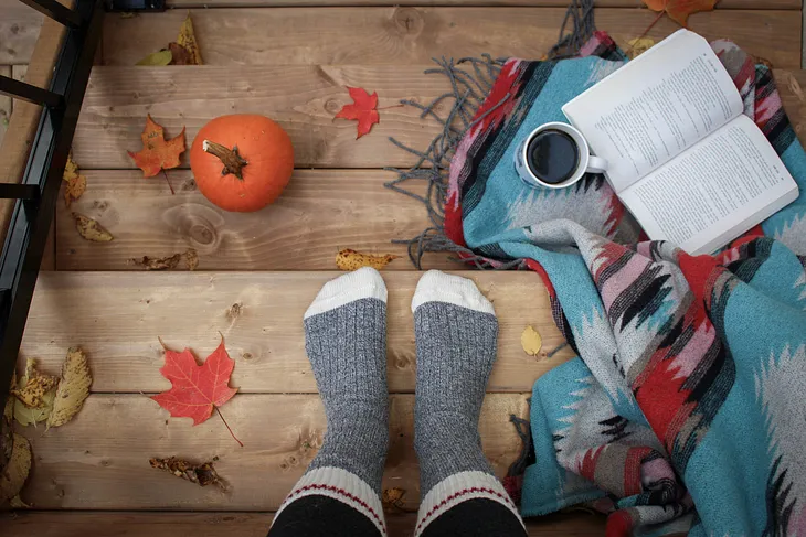 Photo of feet on a wooden steps, with an open book beside them, and a cup of black coffee.