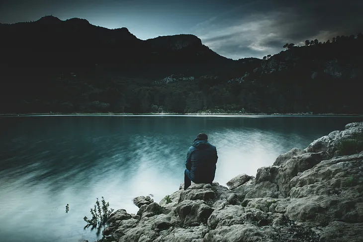 A man sitting buy a lake with his back turned and staring at the lake and the hills in the background.
