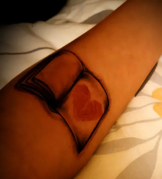 A light brown arm featuring a tattoo of a book that is open to the last page. On the last page are a set of red fingerprints in the shape of a heart.