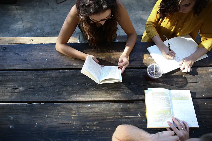 Two people reading and another person writing on a picnic table.