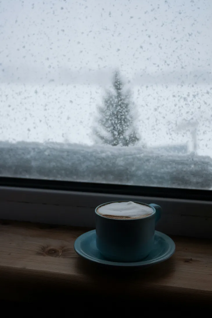 A mug is full to the brim with frothy coffee. It is placed next to a window which shows a blurry snowy landscape outside.