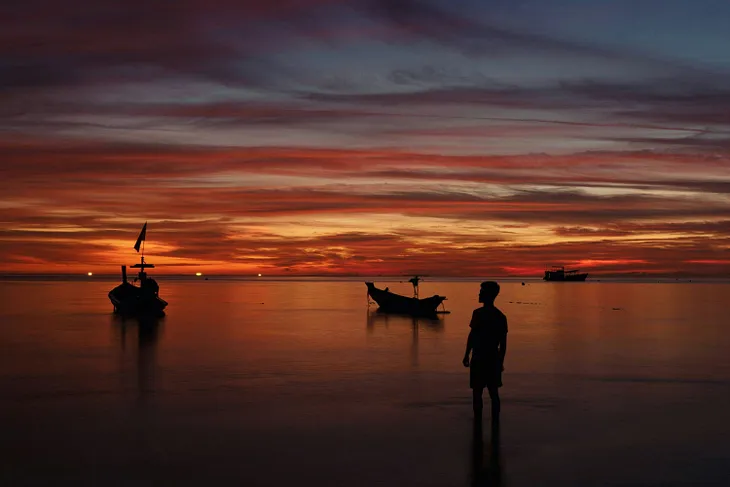 Beautiful sunset in deep orange tune, with a man looking at the horizon. Two little boots near by the man. The presence of Tao is everywhere.