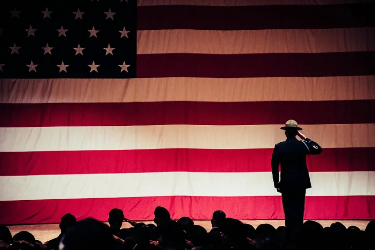 photo of person in US military uniform saulting american flag, standing, in front of seated persons also saluting flag. Posted on MDM Group President Mickey Markoff’s 2024 article on Veterans Day and net worth of selfless sacrifice