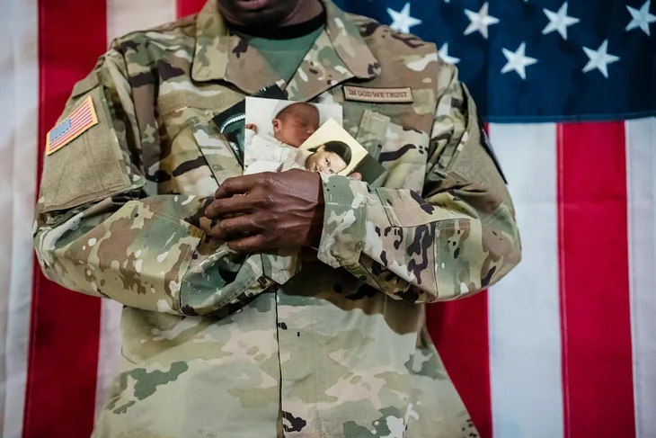 photo of person in camoflauge uniform with american flag in background, holding photos of a baby and a woman, posed on mickey markoff 2024 article on day of the deployed