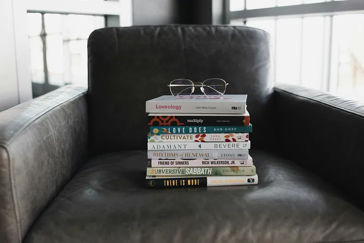 A pile of books on a grey chair with a pair of glasses on top.