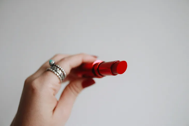 Photograph of a White hand holding a bright red uncovered lipstick, towards the camera.