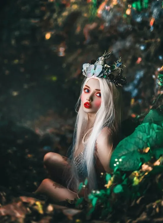 A fantasy creature with long white hair and jewels on her head is looking up over her left shoulder, while hiding on a rocky cliff near water