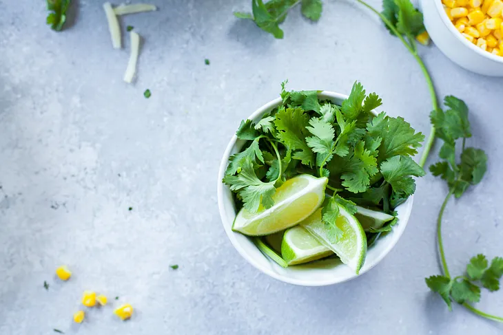 looking down on a white counter with a white bowl of cilantro leaves and a couple wedges of lime