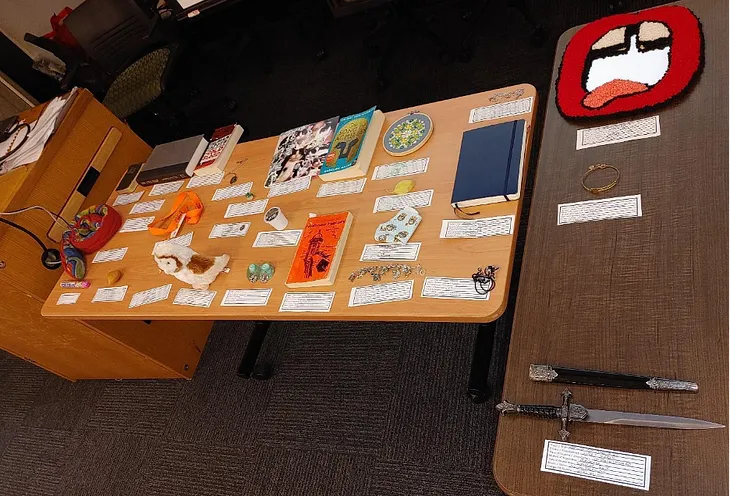 Image 1: Two rectangular tables with personal belongings placed on top, accompanied by hand written labels. Some of the belongings include a packet of Mentos, a stuffed toy snake, books, a cat collage, jewellery, a stuffed toy owl, and a dagger.