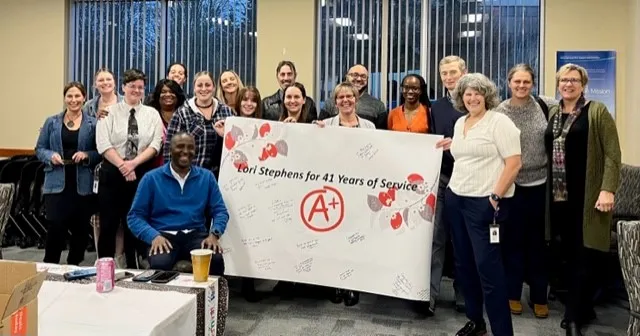 A group of nearly 20 people are gathered around a banner that says “Lori Stephens for 41 Years of Service” and “A+” on it. They are standing inside a room in front of windows. The people are smiling.