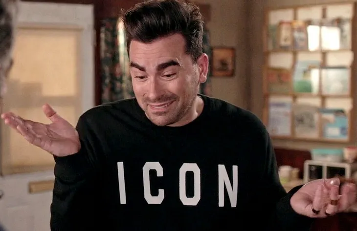 David Rose from Schitt’s Creek wearing a black sweater that reads “icon” in large capital white letters, his hands held up, a slight smile on his face.