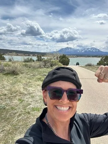 CC dressed in a black long sleeve shirt and black cap with sunglasses smiling and doing a strong arm selfiie in the camera with a background of mountains and a lake.