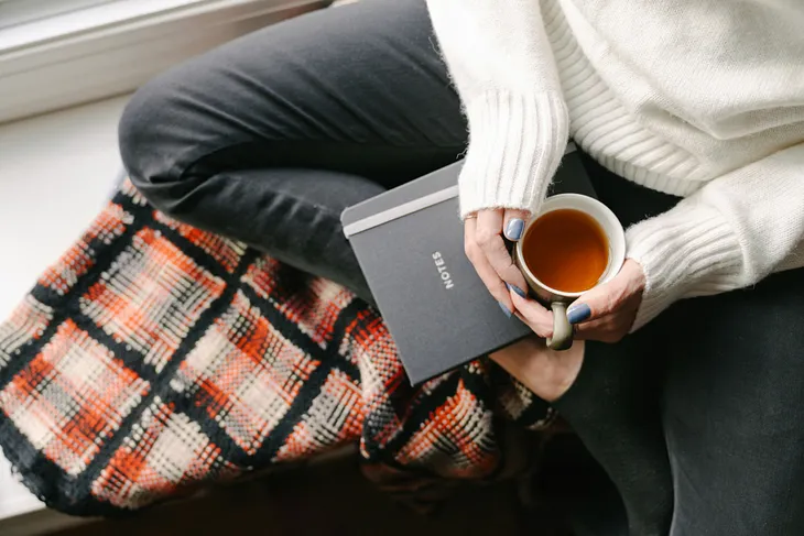 Woman wearing white sweater and jeans sitting on windowsill with notebook and cup of tea, on top of cozy blanket.