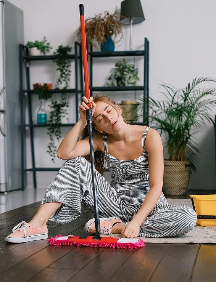 A lady sitting on the floor is fast asleep with a mop, and she looks like she had fallen asleep while doing some house chores. In the background is a wall unit with decorations and hanging plants, and beside it is a one-potted indoor palm tree. She is wearing a thin strap, sleeveless overalls, and casual shoes.