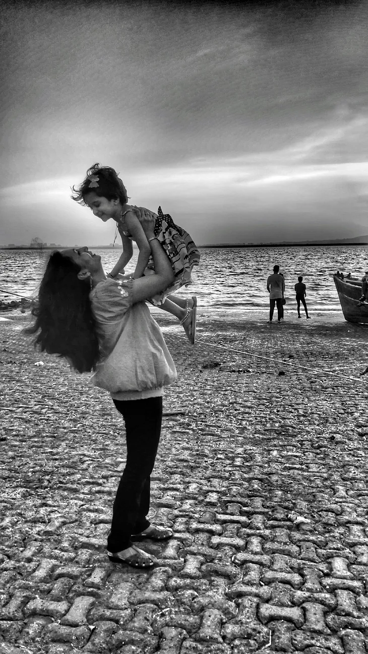 A woman holds a young girl up in the air, at a beach or harbour