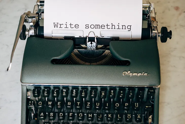 Old school typewriter on a desk with a piece of paper rolled into it. The paper reads: “write something”