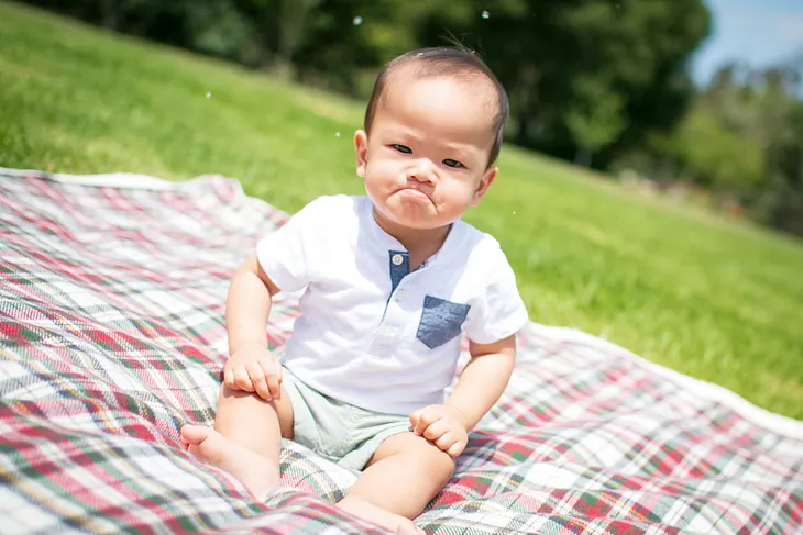 The image shows a young child sitting on a plaid blanket outdoors. The child has a serious, almost pouty expression on their face, with furrowed brows, as they sit on a grassy area with blurred greenery in the background.