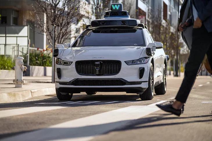 IMAGE: A photo of a Waymo Jaguar iPACE in an urban setting, with a pedestrian crossing in front of it