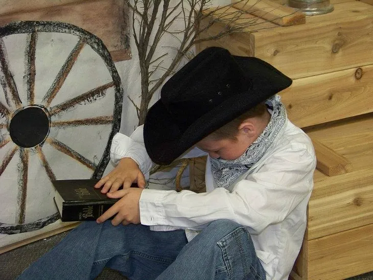 A small boy in blue jeans, white long-sleeved shirt, white and black bandana around his neck and black cowboy hat sitting against two wooden crates by a small branch and wagon wheel drawn on paper against the wall with his head bowed and his right hand on a Bible that he is holding with his left hand.