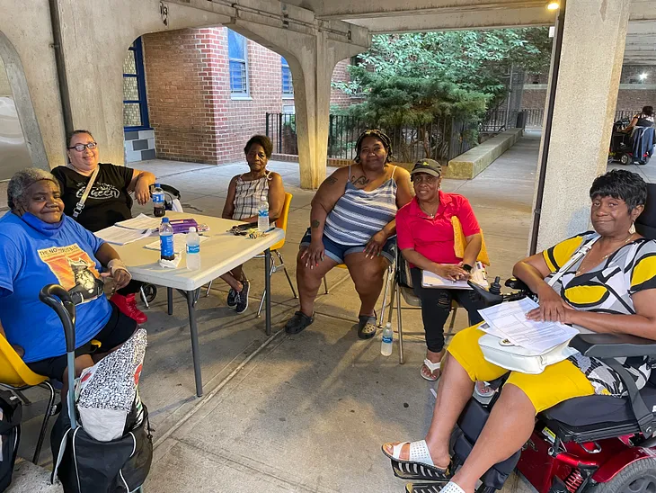 Six women sitting around a table outside an apartment building looking at the camera.