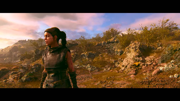 Hellblade II’s hero Senua stands in front of some rocks and grass, under a soft sun-lit sky.