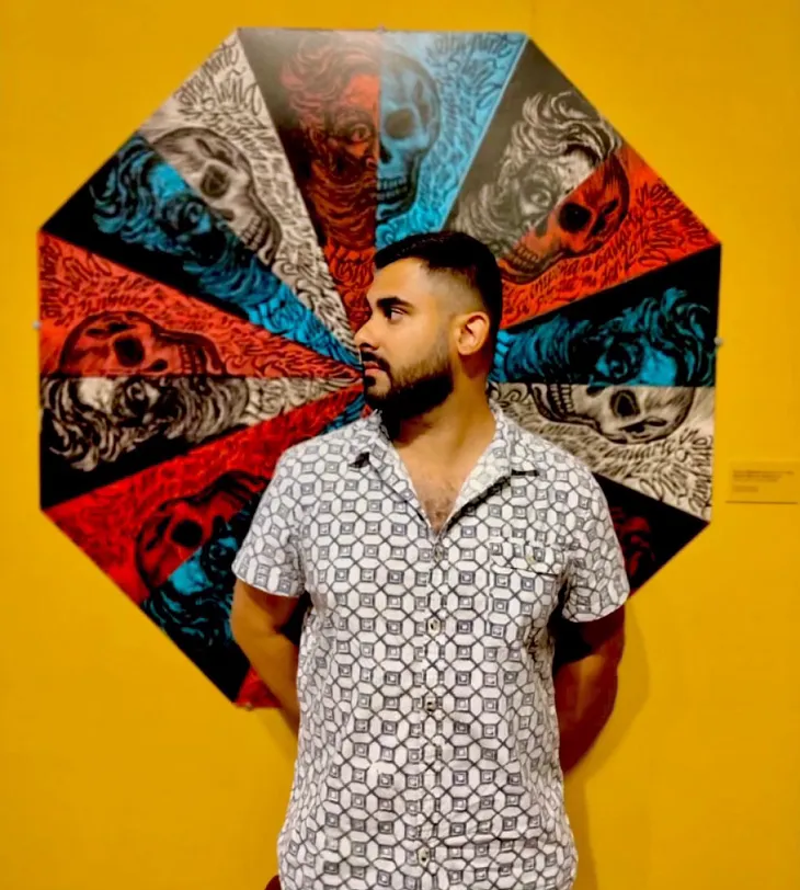 Alt text: A man with short dark hair and a beard stands in front of a colorful piece of circular artwork featuring alternating patterns of red, blue, and black skull designs. He is wearing a light-colored patterned short-sleeve button-up shirt, looking to his left with his hands behind his back. The background is a mustard yellow wall, and the artwork behind him forms an umbrella-like shape.