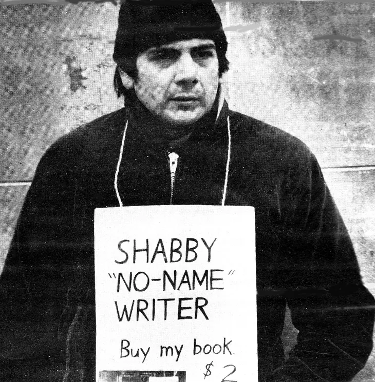 A black and white picture of Crad Kilodney — a middle-aged, gruff-looking white man in his 30s — in a toque and winter coat, wearing a sign around his neck that reads ‘Shabby ‘No-Name’ Writer — Buy my book — $2.’