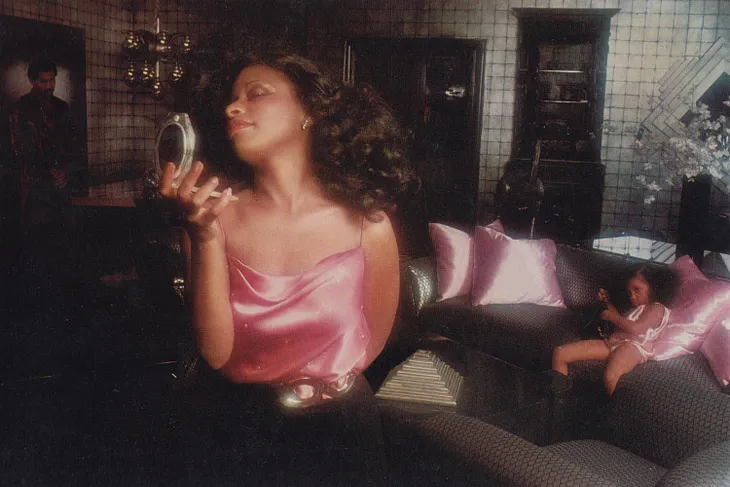 This wide-framed image of Chaka Khan depicts her in a living room (poshly decorated for the 1980s). She gazes at herself in a compact mirror while wearing a spaghetti-strapped pink camisol top with her waist cinched by a black belt with a dark gray buckle around hip-hugging black pants. Of to her left in shadow is a swarthy gentleman gazing at Chaka. Over Chaka’s right shoulder, we see her daughter Milini in pink pajamas looking at her mom getting ready for a night out on the town.