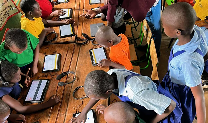 A group of kids wearing colorful shirts, gather around a table, looking at and engaging with their tablets, learning.